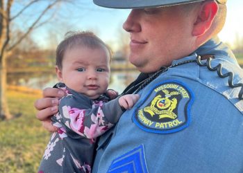 A Missouri State Highway Patrol officer holds a baby in floral clothing outdoors on a sunny day.